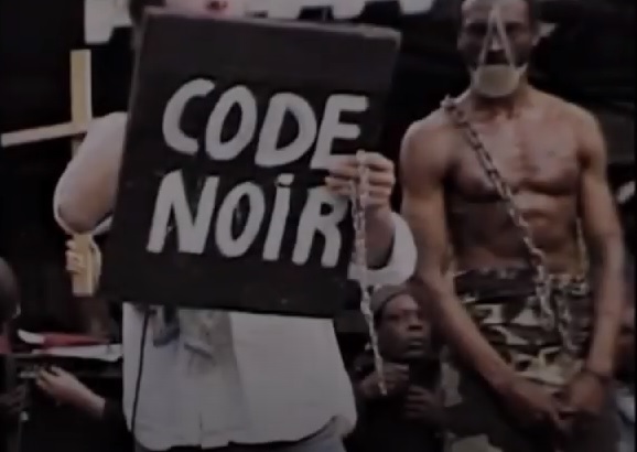 Plan théâtral rapproché qui décrit une vente d' esclaves, Nantes 2006. Un trafiquant brandit la pancarte sur laquelle est inscrit CODE NOIR, à côté d'un africain enchaîné et ballonné.